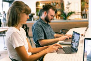 A seated woman at a coffeehouse works on her laptop while a man seated next to her uses his laptop.